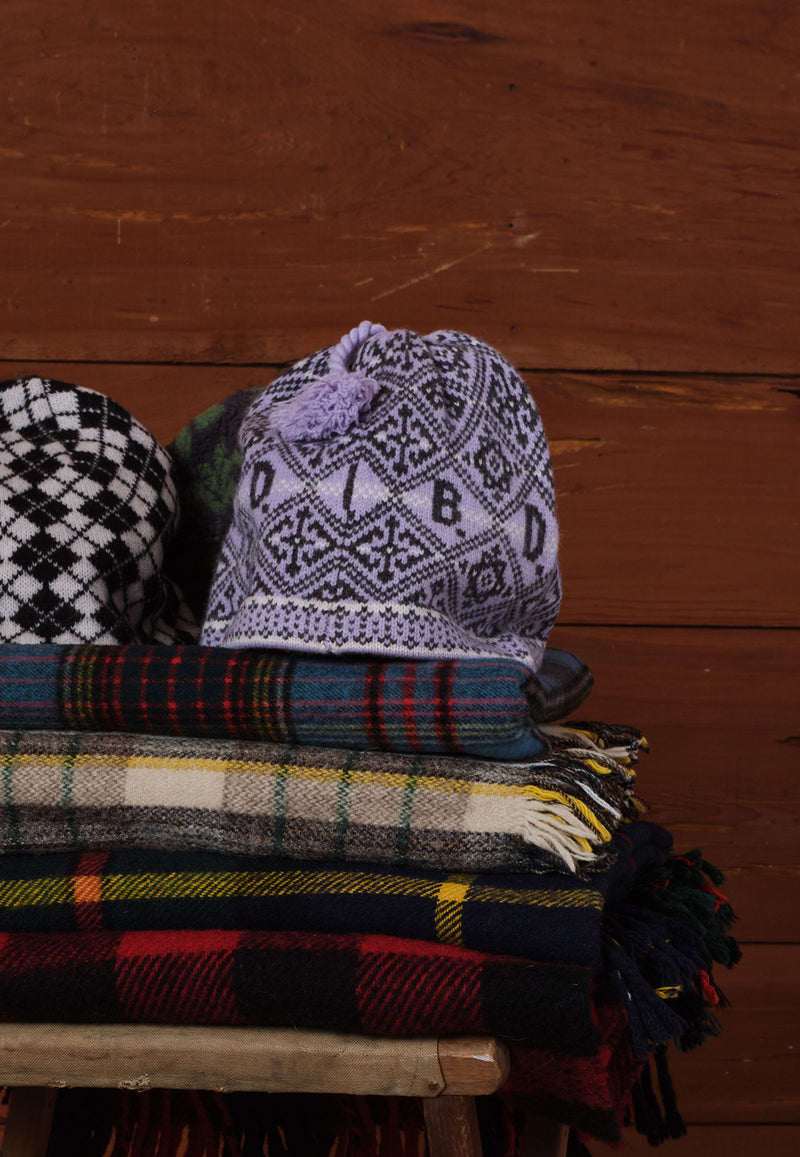 A knitted beanie hat with a purple pom-pom and black geometric pattern sits on top of folded plaid scarves and blankets against a rustic wooden plank 