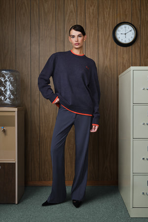 A model stands in an office-like room with wood-paneled walls, wearing a dark grey sweater and matching wide-leg trousers with bright orange trim details.
|M1