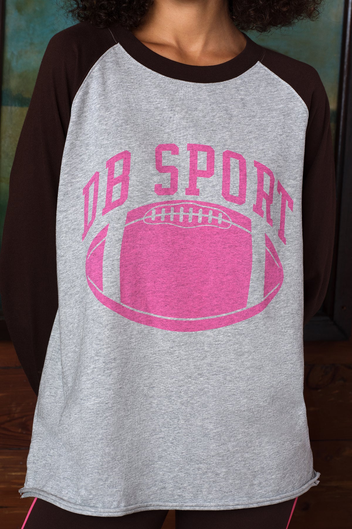 A close-up of a person wearing a heather gray and dark brown raglan t-shirt with a pink graphic that reads 'DB SPORT' above a pink football.
|M1