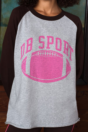A close-up of a person wearing a heather gray and dark brown raglan t-shirt with a pink graphic that reads 'DB SPORT' above a pink football.
|M1