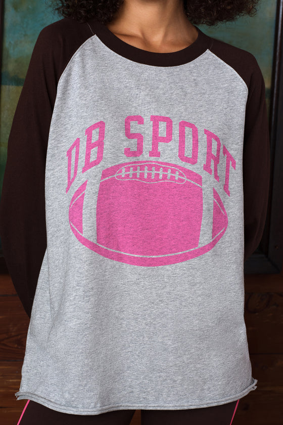 A close-up of a person wearing a heather gray and dark brown raglan t-shirt with a pink graphic that reads 'DB SPORT' above a pink football.
|M1