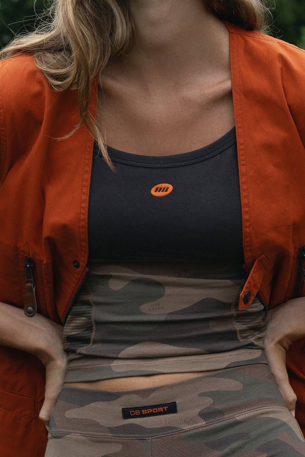 A close-up lifestyle shot of a woman wearing a dark gray tank top with an orange circular logo, a rust-orange zip-up jacket, and camouflage athletic leggings with an orange 'DB SPORT' label.