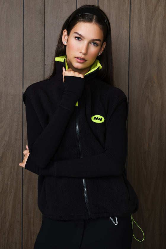 A woman wearing a black zip-up vest with a bright neon yellow-green collar lining and a matching neon yellow-green circular logo patch on the chest, posed against a dark wood-paneled wall.
|M1