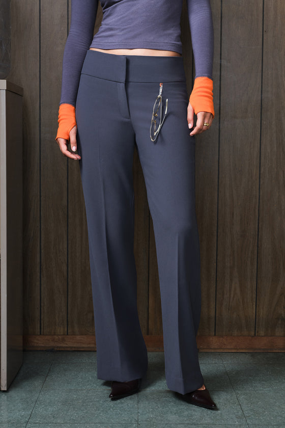 A person wearing high-waisted, wide-leg grey trousers with a matching grey top featuring orange cuffs, standing against a dark wood-paneled wall.
|M1