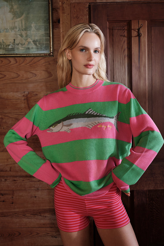 Woman wearing a pink and green striped sweater with a fish design in a wooden interior setting.
|M1