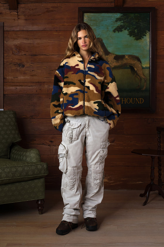 A woman stands in a rustic room with wood-paneled walls, wearing a fuzzy camouflage zip-up jacket and light beige cargo pants.
|M1