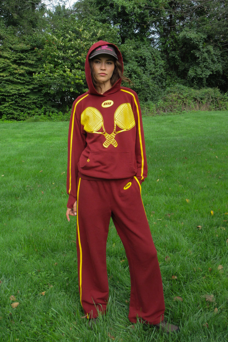 A person standing outdoors on grass wearing a maroon hooded sweatshirt with yellow trim and a large yellow crossed-racket graphic on the front, plus matching maroon pants with yellow side stripes.
|M1