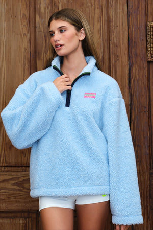 A woman wearing a light blue sherpa fleece quarter-zip pullover sweater with a pink and white logo patch on the chest, posed against a dark wood-paneled wall.
|M1