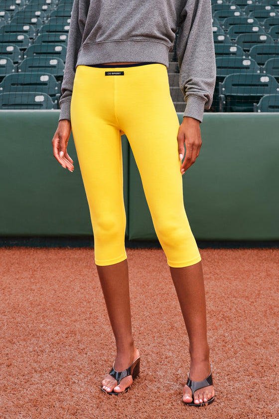 A model wearing bright yellow capri leggings and a gray sweatshirt stands on a baseball field in front of stadium seats.
|M1