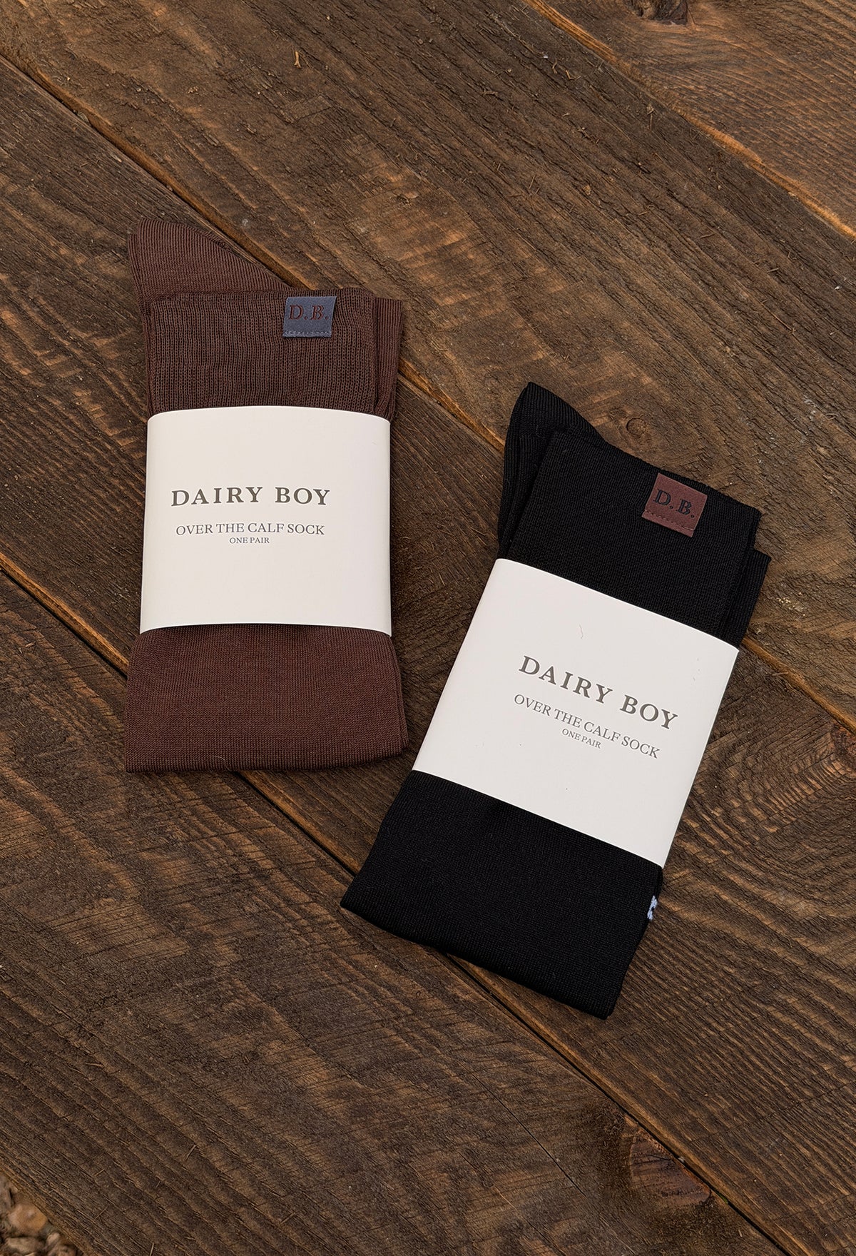 Two pairs of folded socks in brown and black, each wrapped in a white paper band labeled 'DAIRY BOY', placed on a rustic dark wood surface.