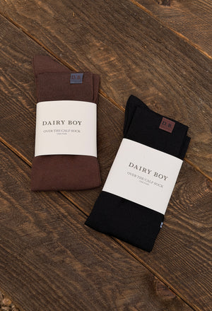 Two pairs of folded socks in brown and black, each wrapped in a white paper band labeled 'DAIRY BOY', placed on a rustic dark wood surface.