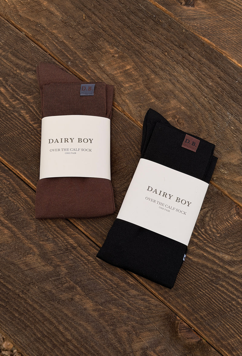 Two pairs of folded socks in brown and black, each wrapped in a white paper band labeled 'DAIRY BOY', placed on a rustic dark wood surface.