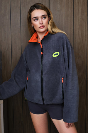 A woman wearing a dark gray fleece zip-up jacket with a bright orange collar and a lime green chest logo, paired with dark grey shorts, standing against a dark wood-paneled wall.
|M1