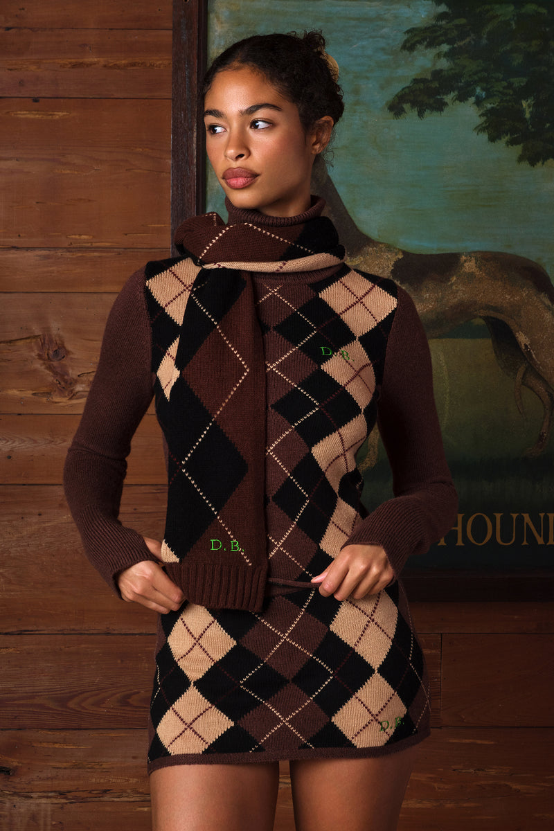 A woman models a brown and beige argyle sweater vest with a matching knit cowl neck scarf. The vest features a classic diamond pattern in dark brown, medium brown, and tan, with small green embroidered initials 'DB.
