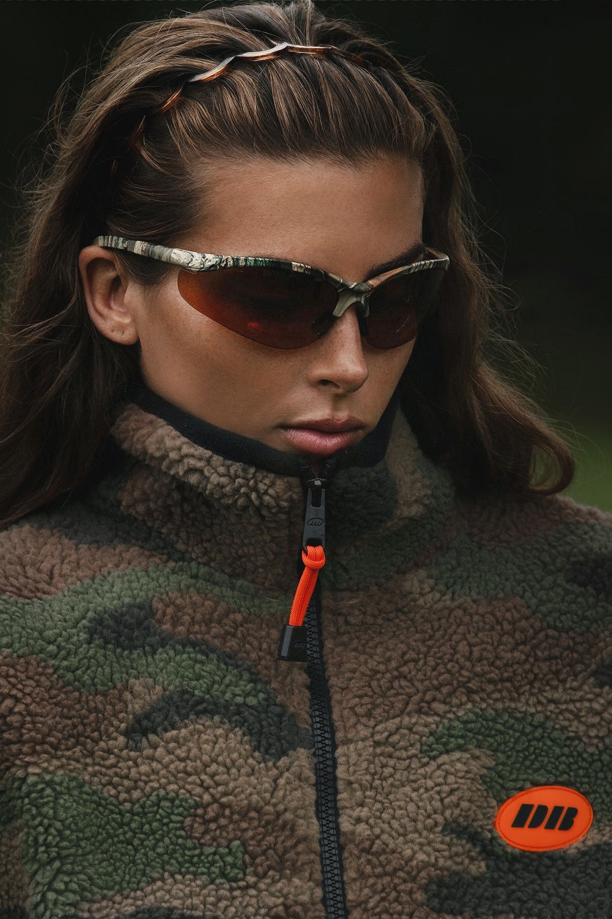 A close-up of a woman wearing camouflage-patterned sunglasses with orange-tinted lenses and a matching camouflage fleece jacket with an orange logo patch on the chest.
|M2
