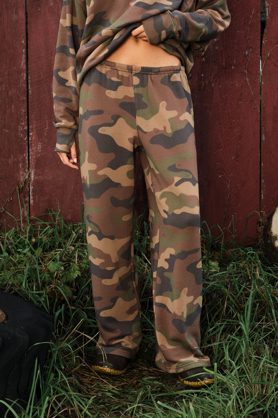 A person wearing loose camouflage sweatpants and a matching camouflage top, standing outdoors in front of a red wooden fence and grass.
|M1