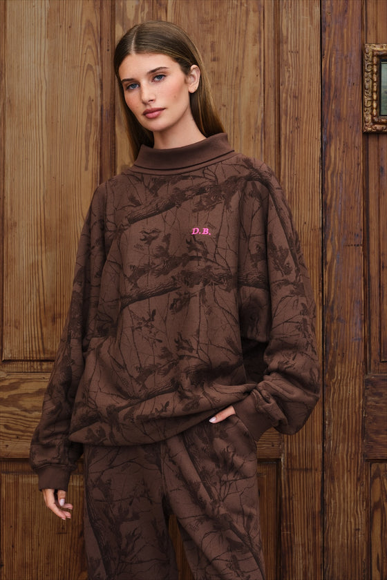 Model stands wearing brown camo rollneck sweatshirt with pink "D.B" monogram logo on the chest, in from of a rustic wood paneled wall. 
|M1