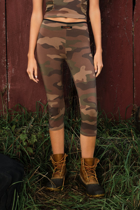 A model wearing camouflage print capri leggings and a matching camouflage top, standing outdoors in front of a rustic red wooden wall and green grass.
|M1