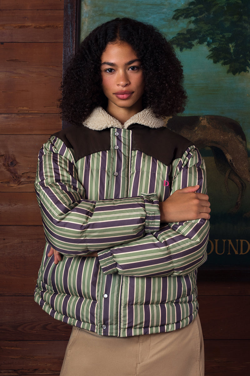 A model with curly dark hair wearing a striped puffer jacket with a brown yoke, cream sherpa collar, and green/brown vertical stripes, posed against a rustic wood-paneled wall.
|M1