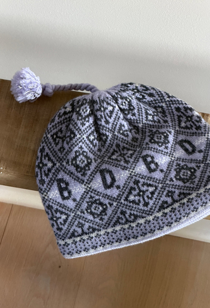 A knit lavender beanie hat with a gray and white geometric pattern featuring the letters 'DB' and a matching purple tassel, resting on a wooden surface.