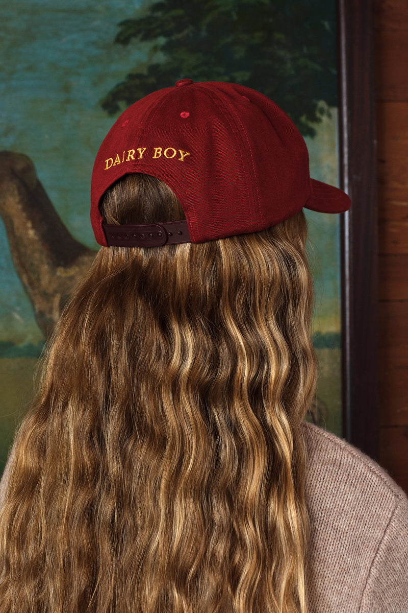 A person with long wavy light brown hair seen from the back, wearing a dark red baseball cap with 'DAIRY BOY' embroidered in gold on the side. The photo is cropped from the shoulders up against an indoor background with a painting and dark wood paneling.