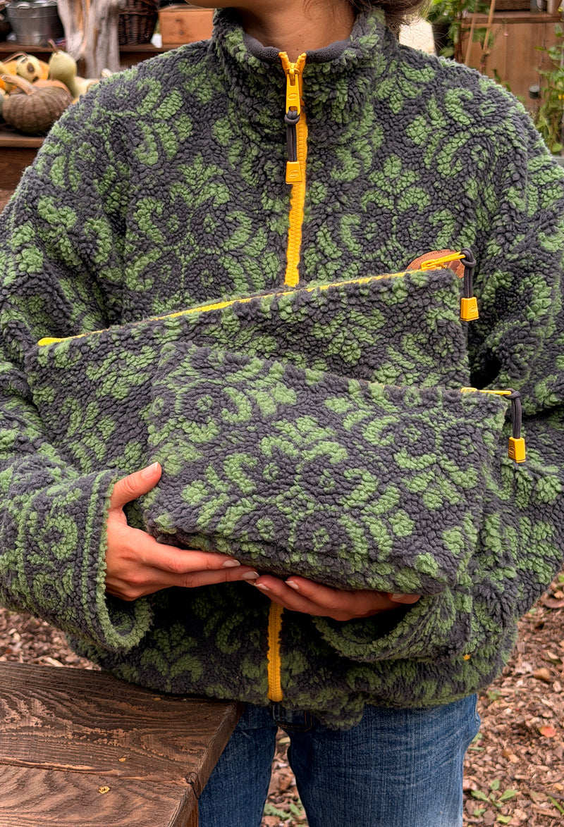A person wearing a dark gray and green patterned fleece jacket holds a matching zippered pouch in front of their torso. The jacket and pouch feature a textured leaf-like pattern in green and dark gray, with bright yellow zipper accents.