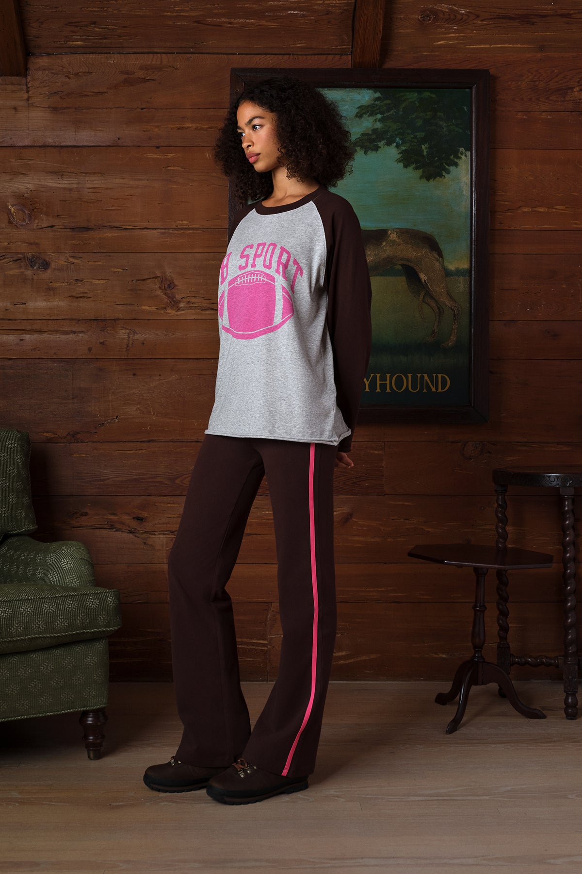 A woman stands in a rustic room with wood-paneled walls, wearing a heather gray and dark brown raglan shirt with a pink football graphic and matching dark brown pants with a pink stripe down the side.
|M1
