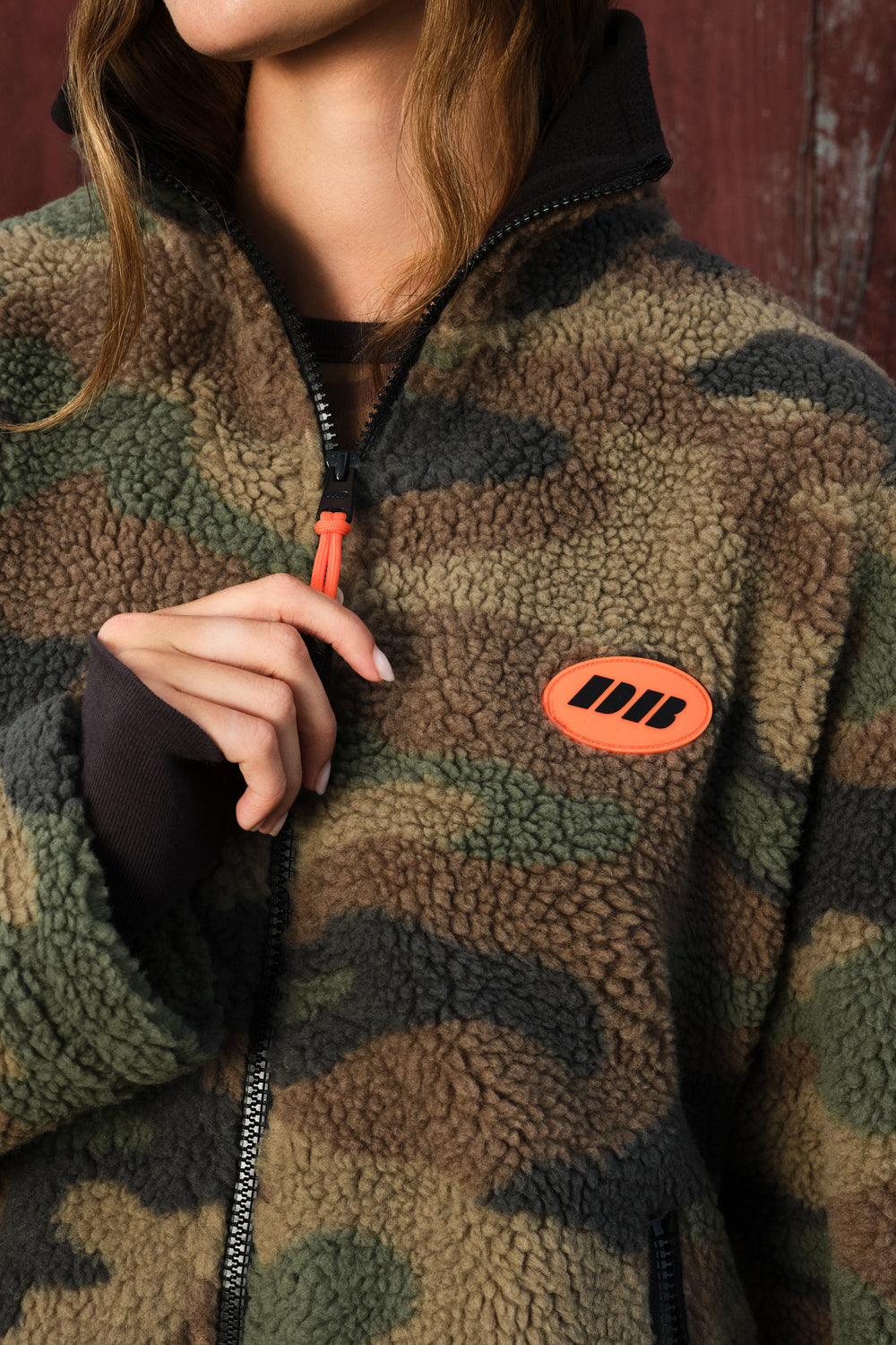 Close-up of a person wearing a sherpa fleece zip-up jacket in a camouflage pattern with a bright orange zipper pull and an orange chest patch logo.
|M1