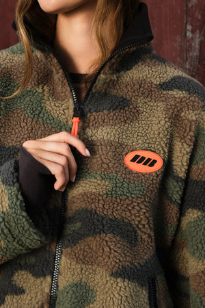 Close-up of a person wearing a sherpa fleece zip-up jacket in a camouflage pattern with a bright orange zipper pull and an orange chest patch logo.
|M1