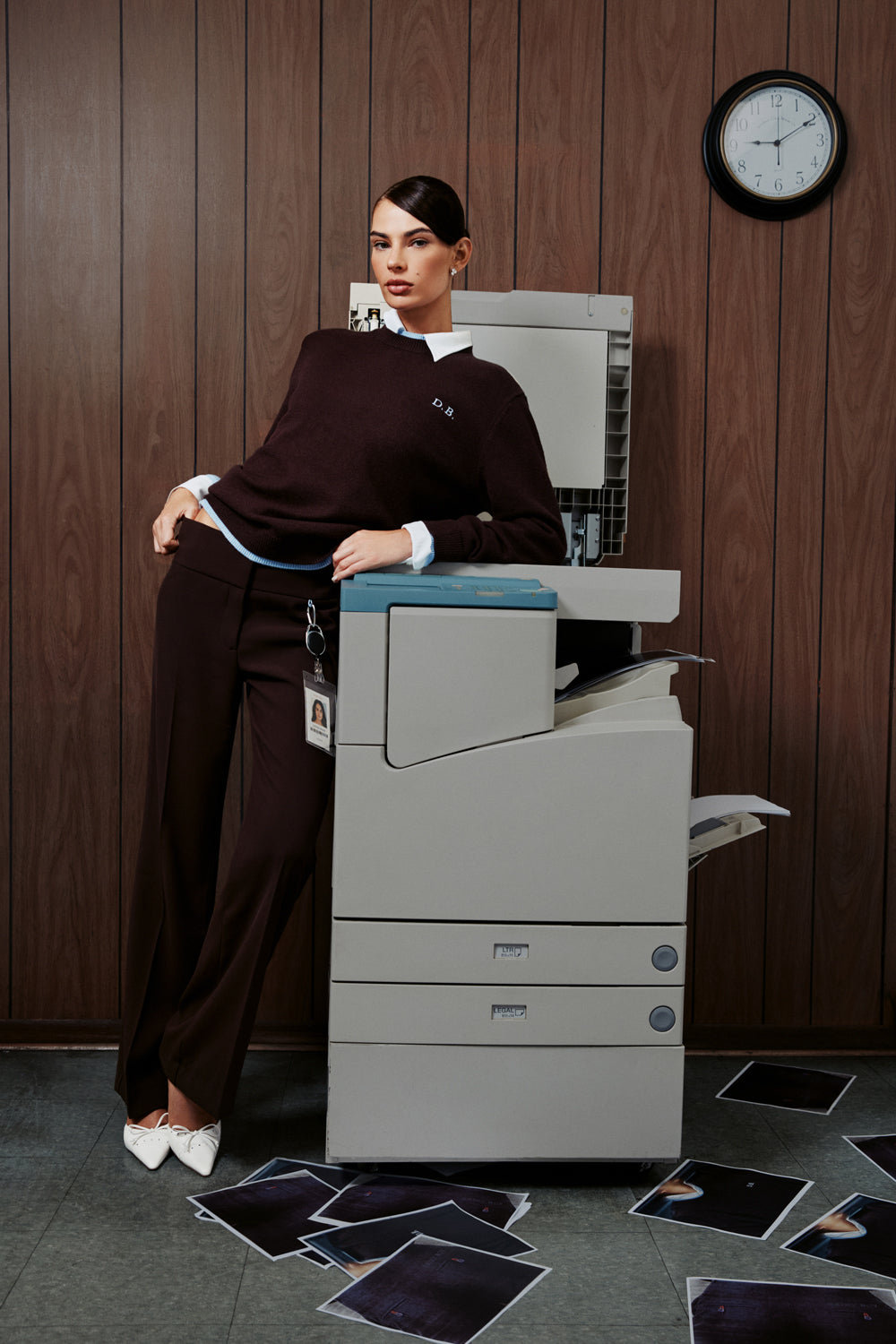 A woman in a dark brown knit sweater and matching wide-leg trousers leans against a large gray photocopier in an office setting with wood-paneled walls, a wall clock, and scattered X-ray film sheets on the floor.
|M1