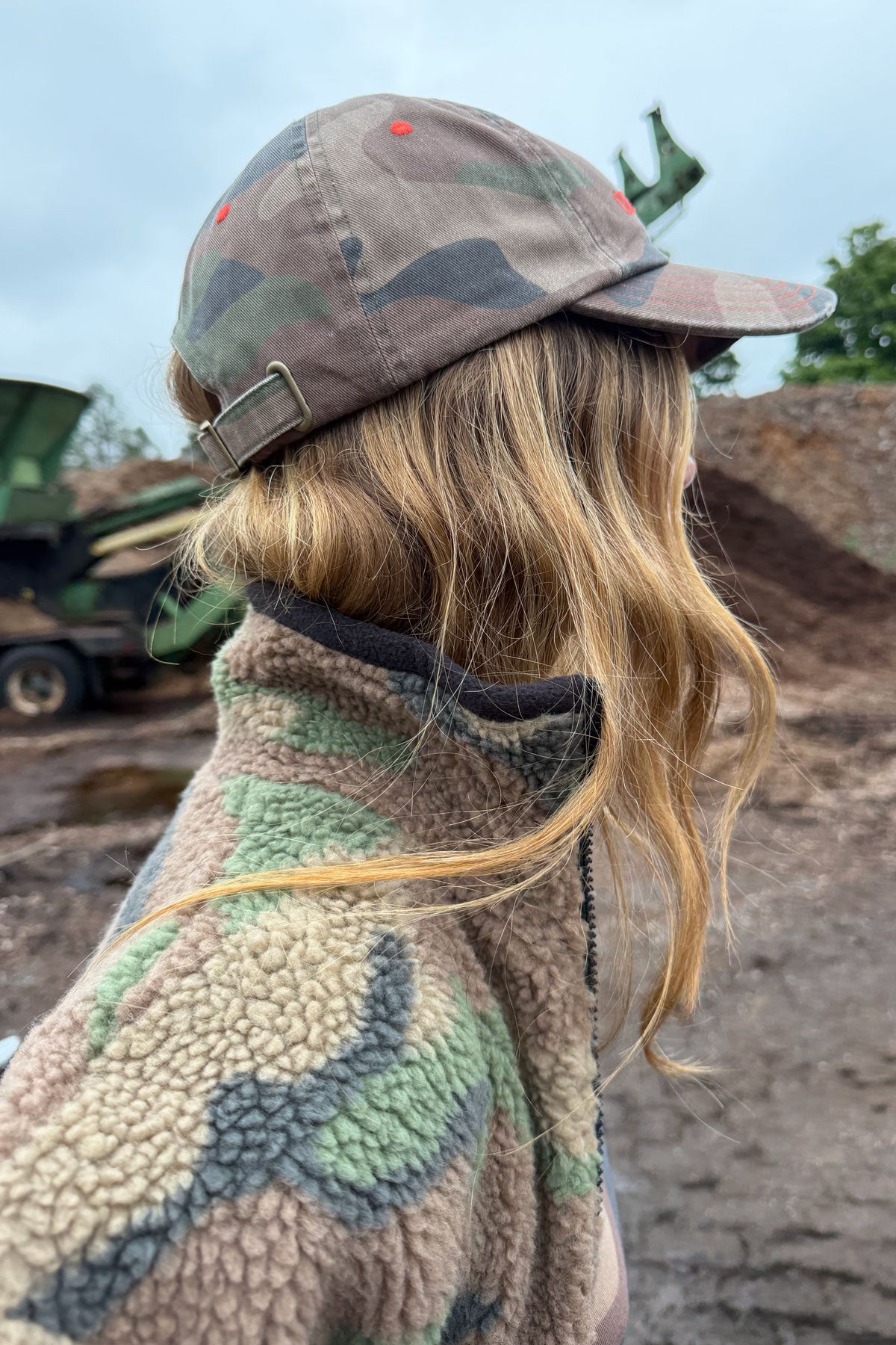 A person shown from the back wearing a camouflage baseball cap and a matching camouflage fleece jacket, standing outdoors in a dusty industrial or construction setting.