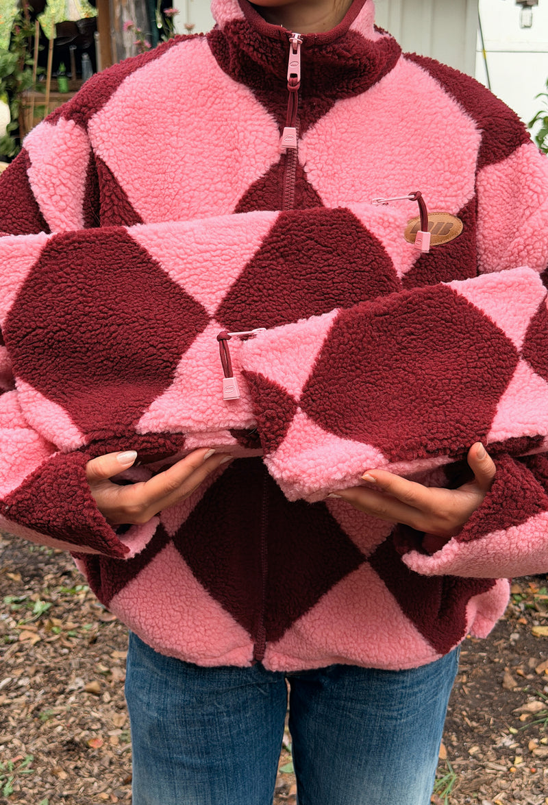 A person wearing a pink and burgundy argyle fleece zip-up jacket, holding two matching fleece pouches with the same argyle pattern. The background is an outdoor setting with trees and ground cover.