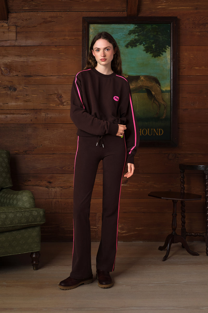 A woman stands in a rustic room with wood-paneled walls wearing a dark brown tracksuit with bright pink piping and a small pink logo on the chest. The set includes a long-sleeve top and matching pants.
|M2