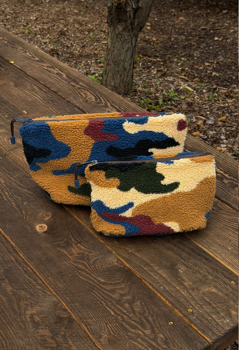Two fleece zip pouches with a multicolor camouflage pattern, resting on a rustic wooden deck outdoors.