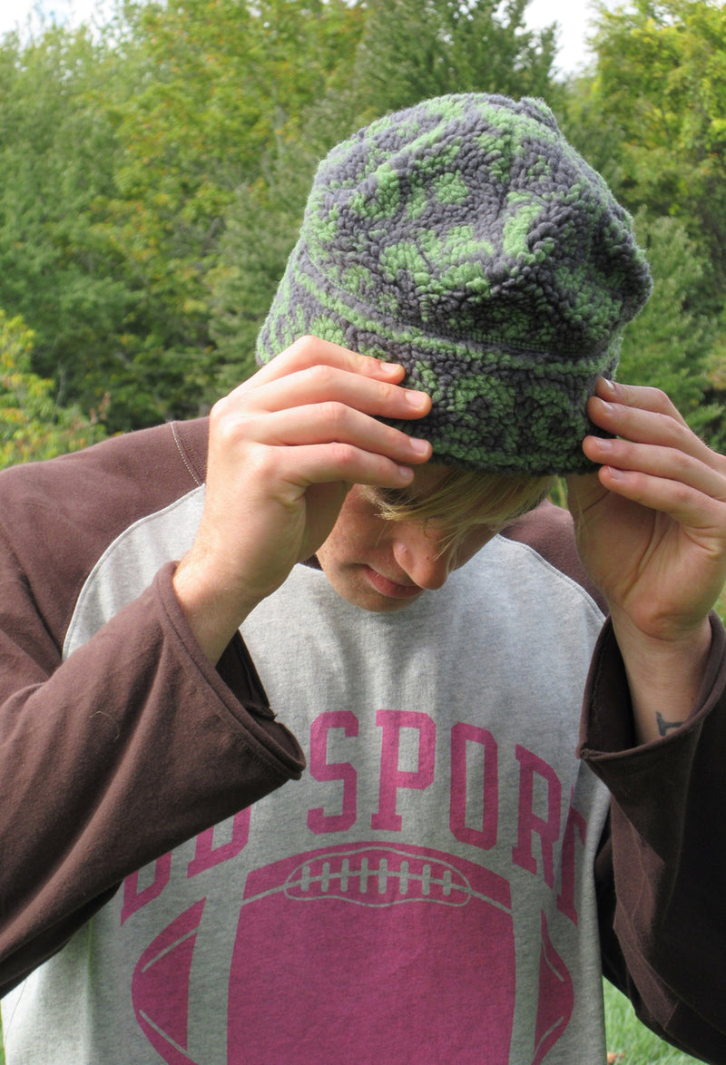 A person outdoors wearing a dark green and gray patterned fleece beanie, holding it in place with both hands. The person is wearing a gray and brown raglan sweatshirt with a pink football graphic.