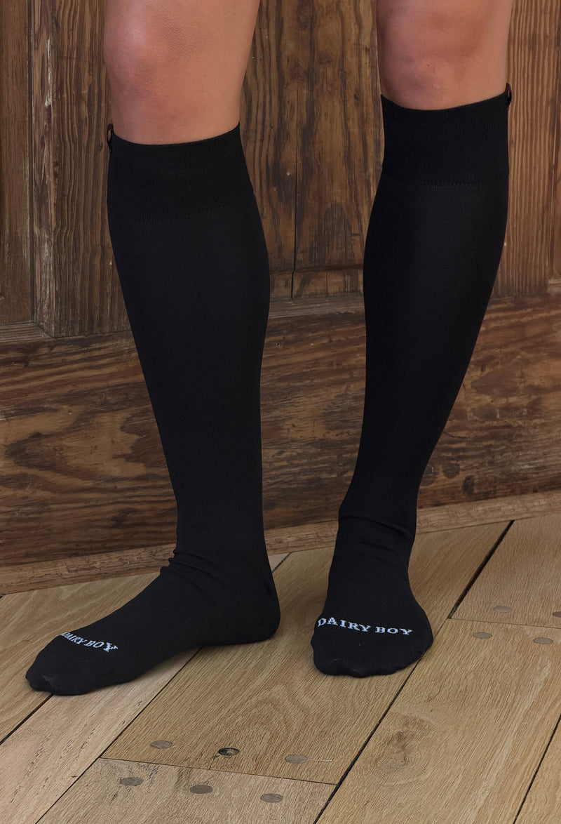 Black knee-high compression socks with white 'DAIRY BOY' branding on the toes, shown on a model's legs against a rustic wooden background.