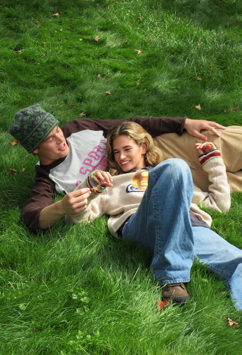 A man and a woman lying on a green grass lawn, smiling and looking at a small object in their hands. The man wears a camouflage beanie, a brown and grey raglan shirt, and tan pants, while the woman wears a beige sweater, blue jeans, and brown shoes.