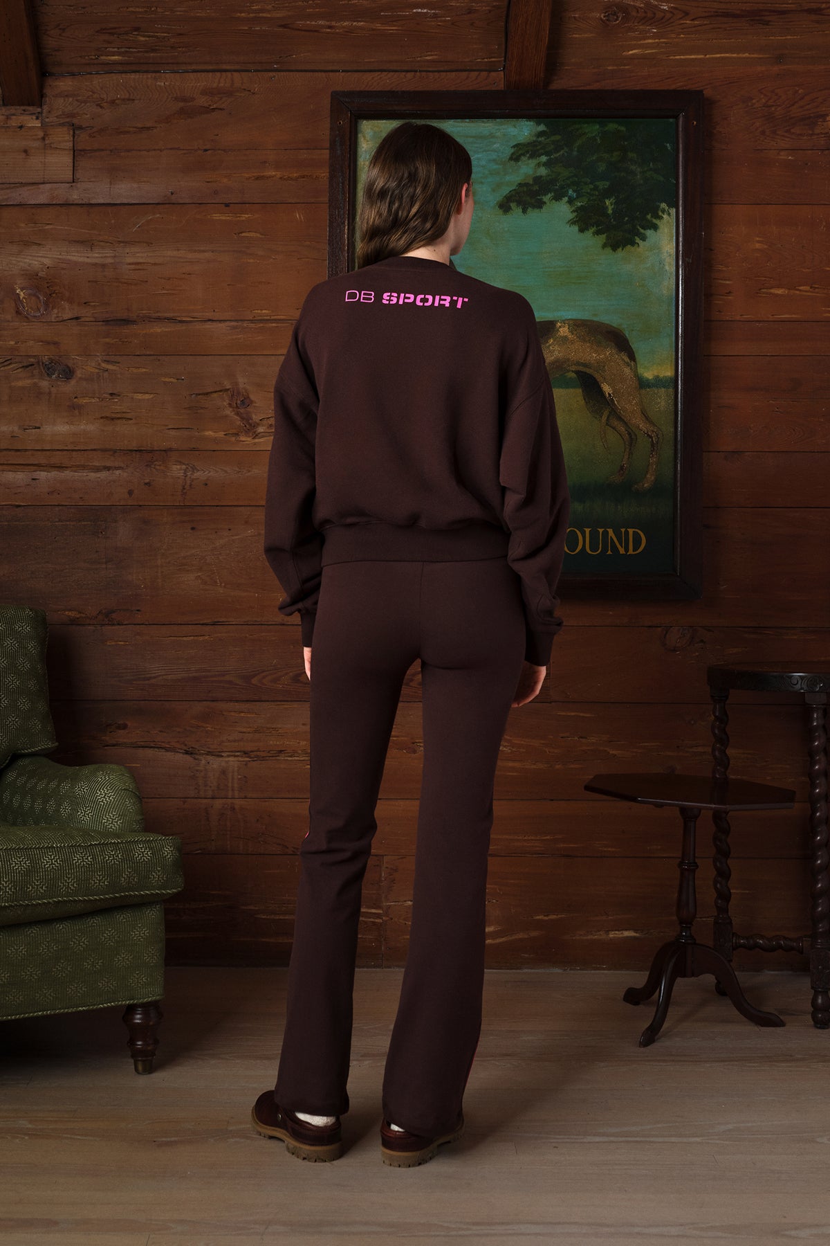 A person stands with their back to the camera in a rustic room with wood-paneled walls, wearing a dark brown sweatshirt and matching pants. The sweatshirt has 'CULT SPORT' printed in pink on the upper back.
|M2