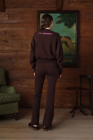 A person stands with their back to the camera in a rustic room with wood-paneled walls, wearing a dark brown sweatshirt and matching pants. The sweatshirt has 'CULT SPORT' printed in pink on the upper back.
|M2