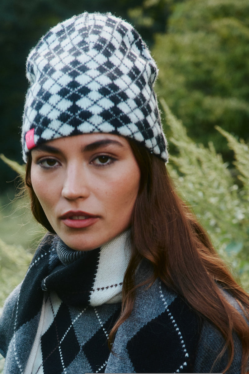 A woman wearing a black and white argyle knit beanie with a small pink tag detail, paired with a matching argyle scarf and gray sweater, set against a soft-focus green outdoor background.