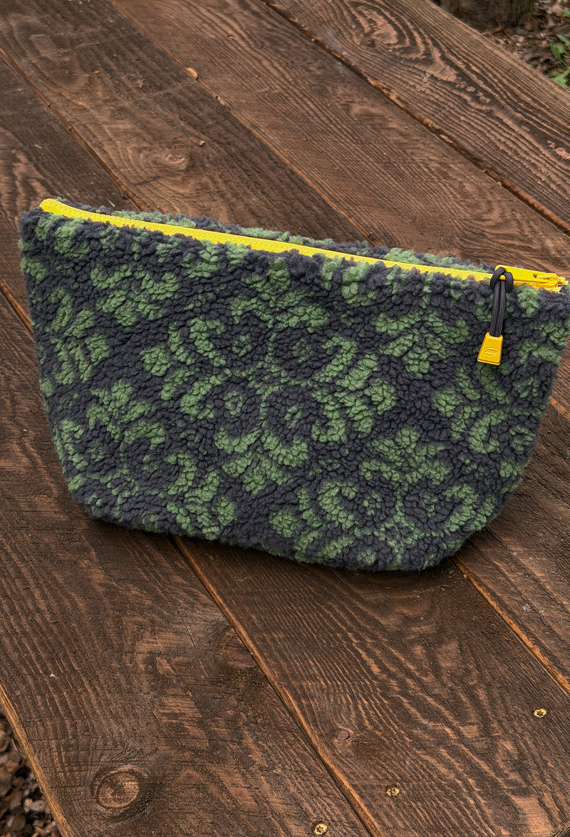 A textured fleece cosmetic pouch with a green and dark gray floral pattern and a bright yellow zipper, resting on a rustic wooden surface.