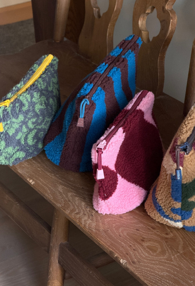Four colorful fleece zippered pouches arranged on a wooden chair. The pouches have distinct patterns and colors: green and gray damask, blue and brown stripes, pink and burgundy argyle, and tan camo with blue and green accents.
