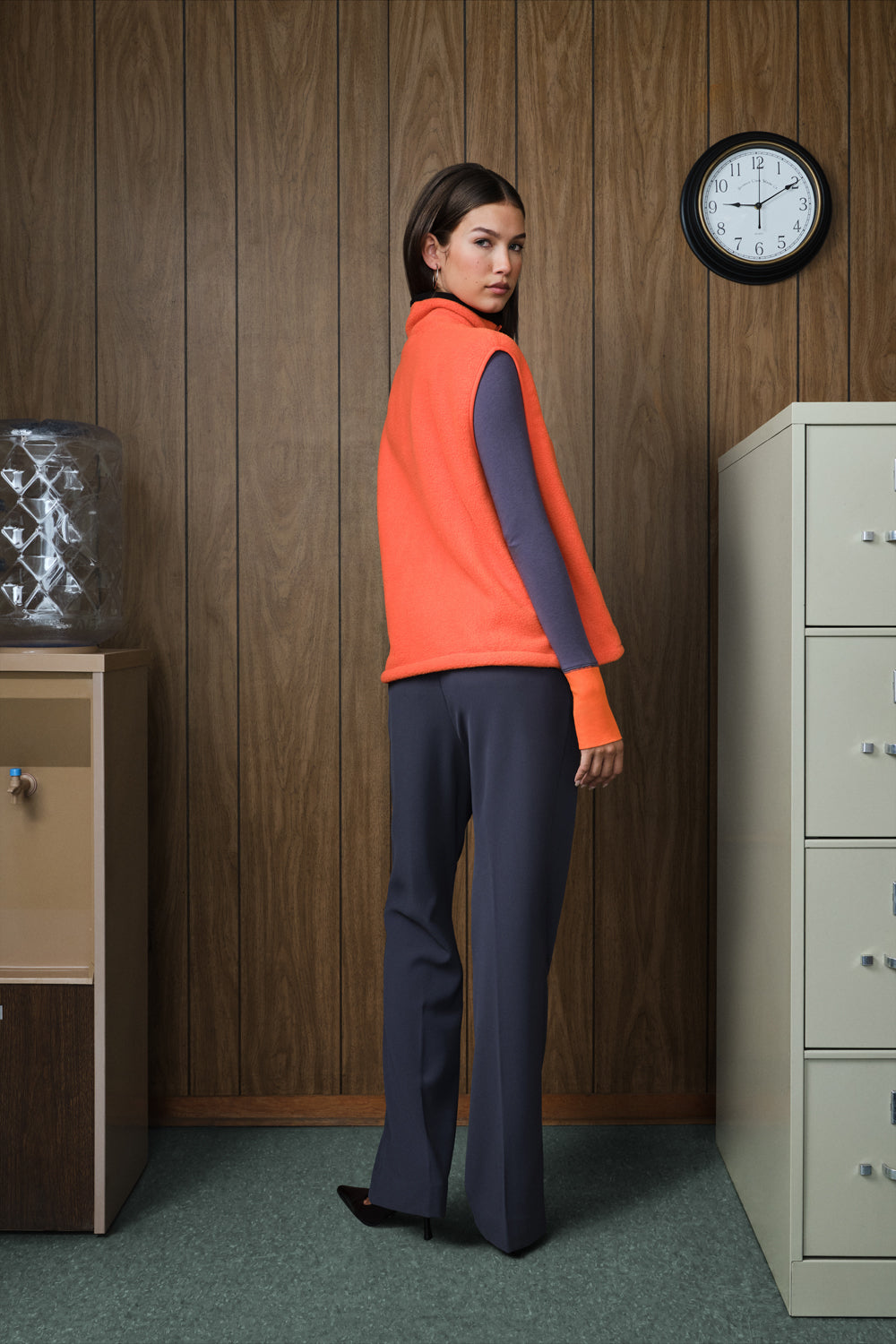 A woman stands in a retro office setting with wood-paneled walls, wearing a bright orange sleeveless vest over a dark grey long-sleeve top and matching grey trousers.
|M1