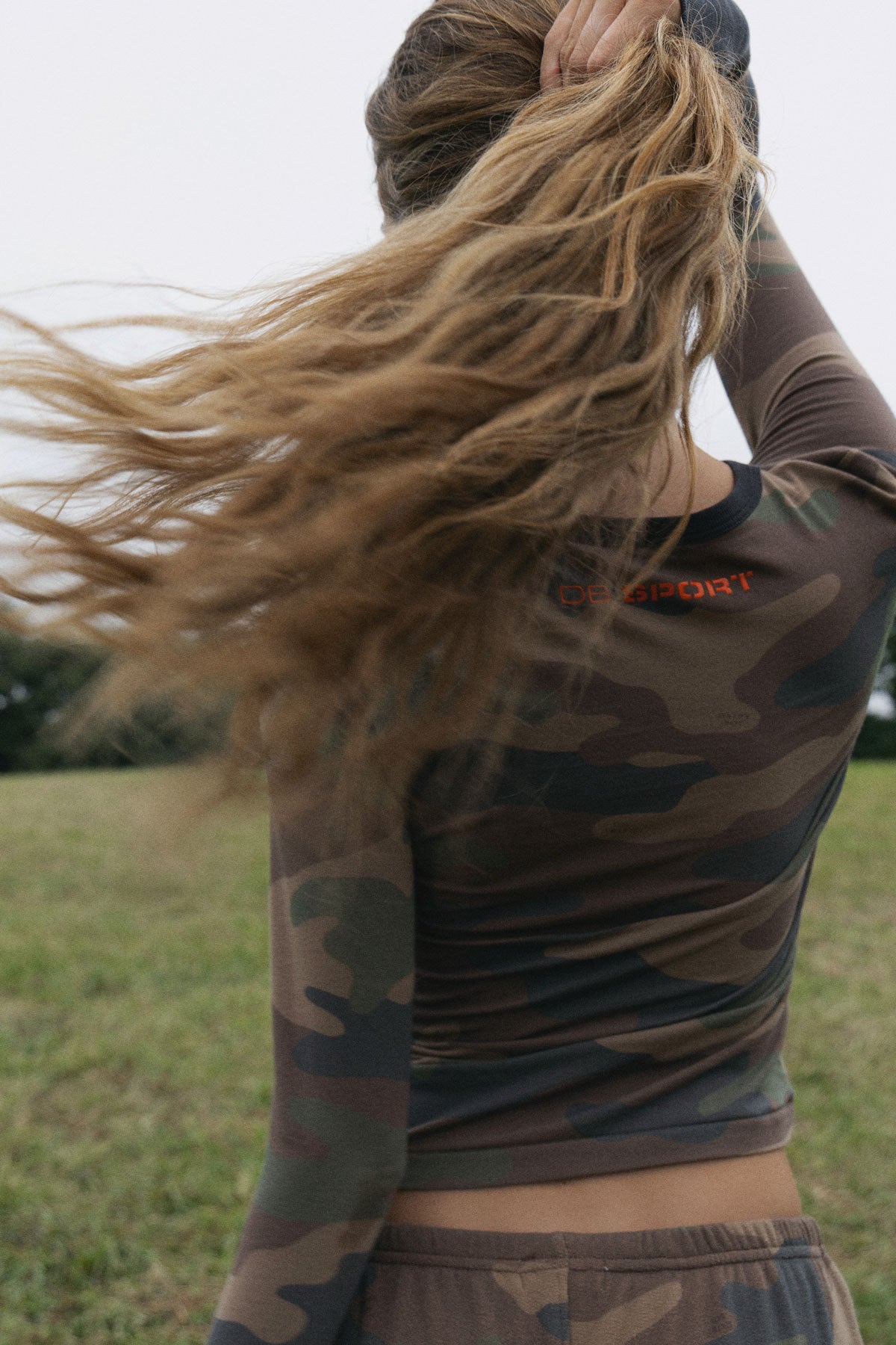 A woman outdoors in a grassy field wearing a fitted long-sleeve camouflage athletic top and matching bottoms, with her hair blowing in the wind. The top has a small orange 'DB SPORT' logo near the upper back.
|M1