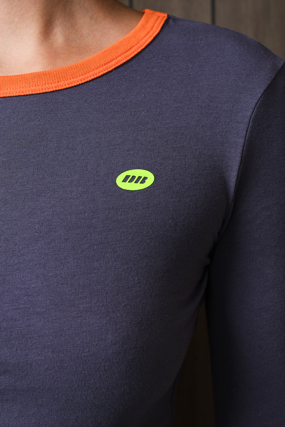 Close-up of a person wearing a dark grey long-sleeve shirt with a bright orange ribbed crew neck and a small lime green oval chest logo.
|M1