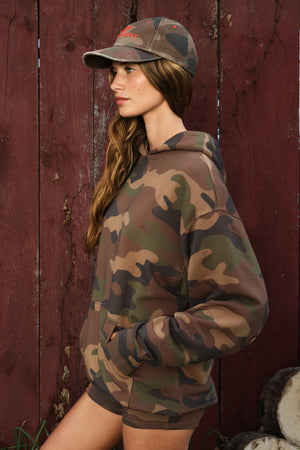 A woman wearing a camouflage hoodie and matching camouflage baseball cap, standing in front of a weathered red wooden wall.
|M1