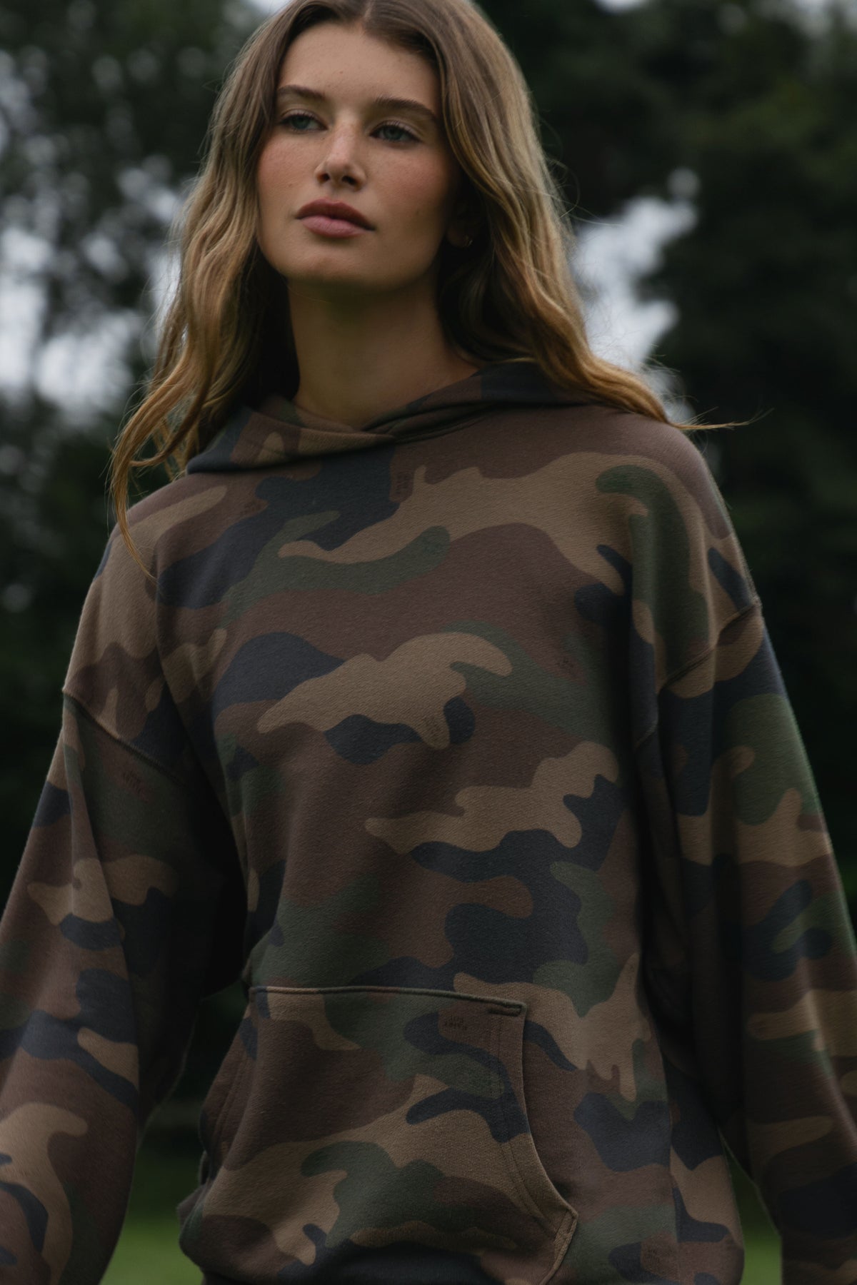 A woman wearing a camouflage hoodie with a front kangaroo pocket, photographed outdoors against a blurred green background.
|M1