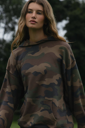 A woman wearing a camouflage hoodie with a front kangaroo pocket, photographed outdoors against a blurred green background.
|M1