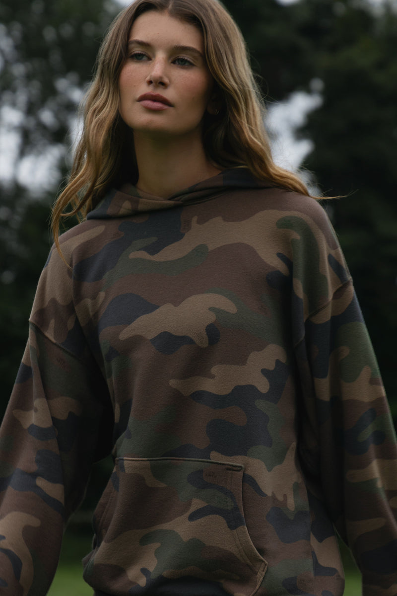A woman wearing a camouflage hoodie with a front kangaroo pocket, photographed outdoors against a blurred green background.
|M1