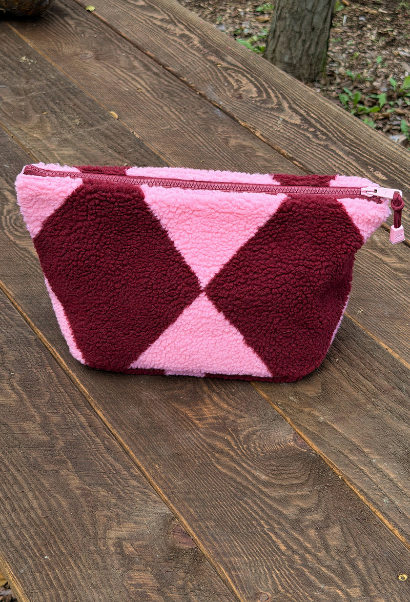 A pink and burgundy fleece zippered pouch with a geometric diamond pattern, resting on a dark wooden plank surface.